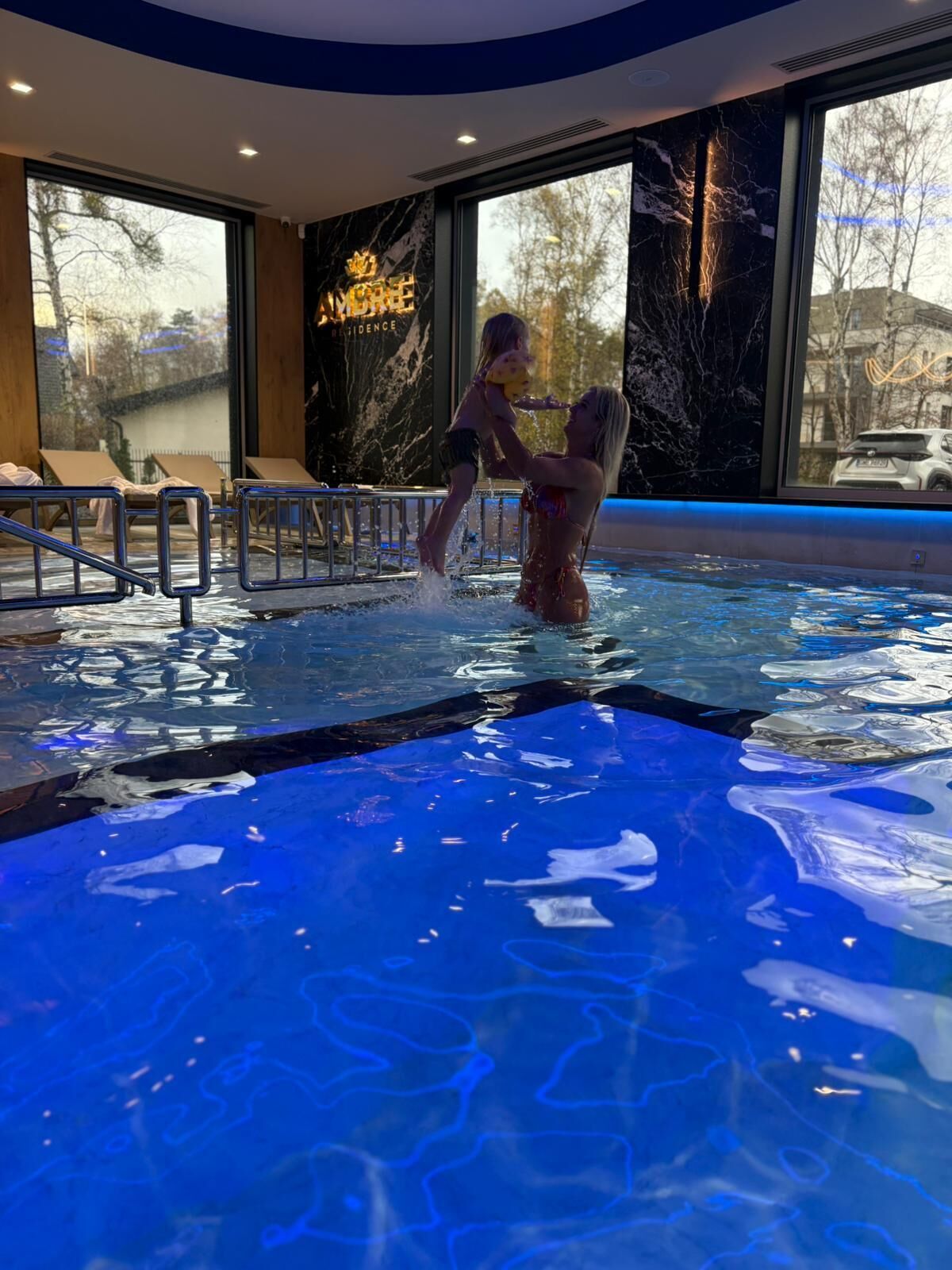 An indoor swimming pool with clear water, a child playing, and lounge chairs.
