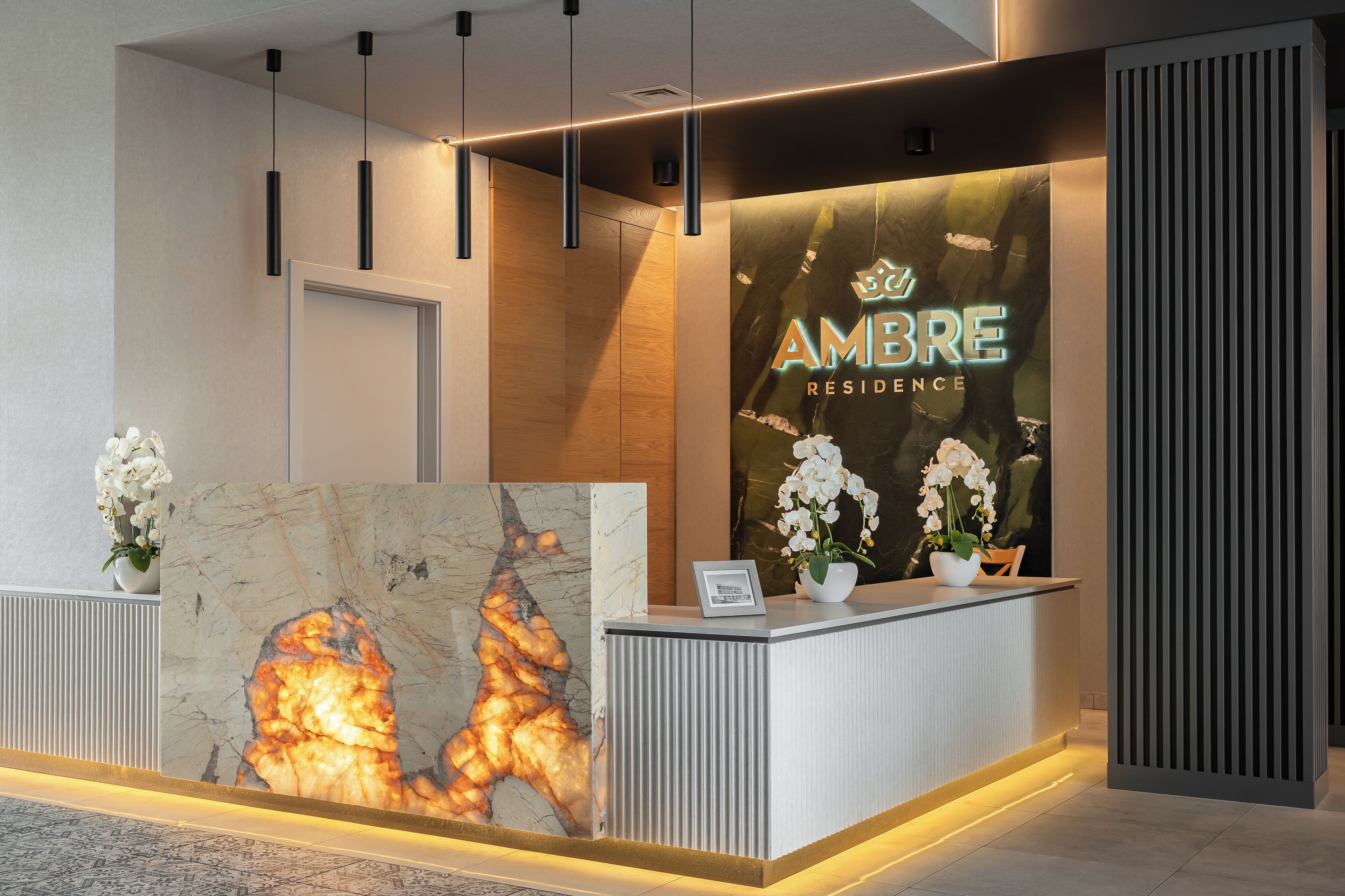 A reception desk with a large illuminated artwork, a sign for 'AMBRE RESIDENCE', and two potted white orchids.