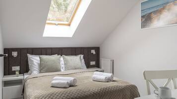 A bedroom with a skylight, a bed with rolled towels, a bedside table, a nightstand, a white chair, and a painting on the wall.