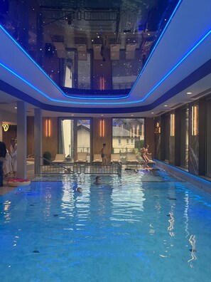 An indoor swimming pool with blue LED lighting, a glass ceiling, and a view of buildings outside. - Rezydencja Ambre (Łeba)