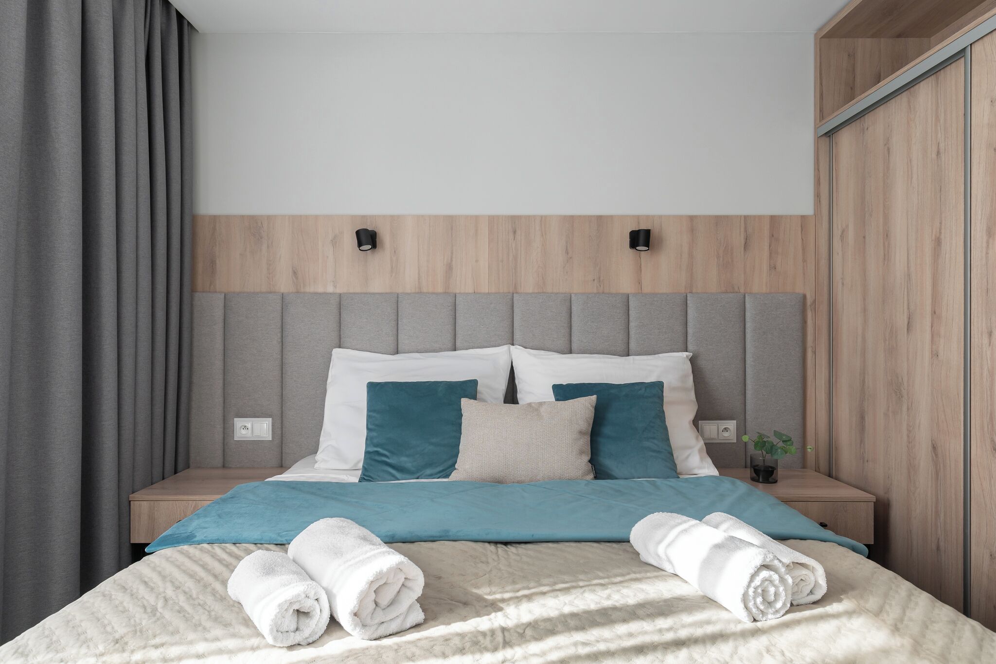 A modern hotel room with a neutral color palette, featuring a bed with neatly folded towels, a wooden headboard, and a small bedside table with a plant.