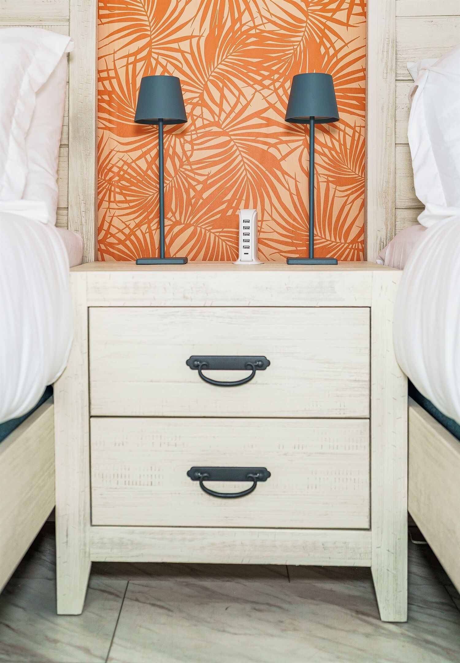 A bedroom with two beds, a nightstand with drawers, a lamp, and a wall with a leaf pattern.