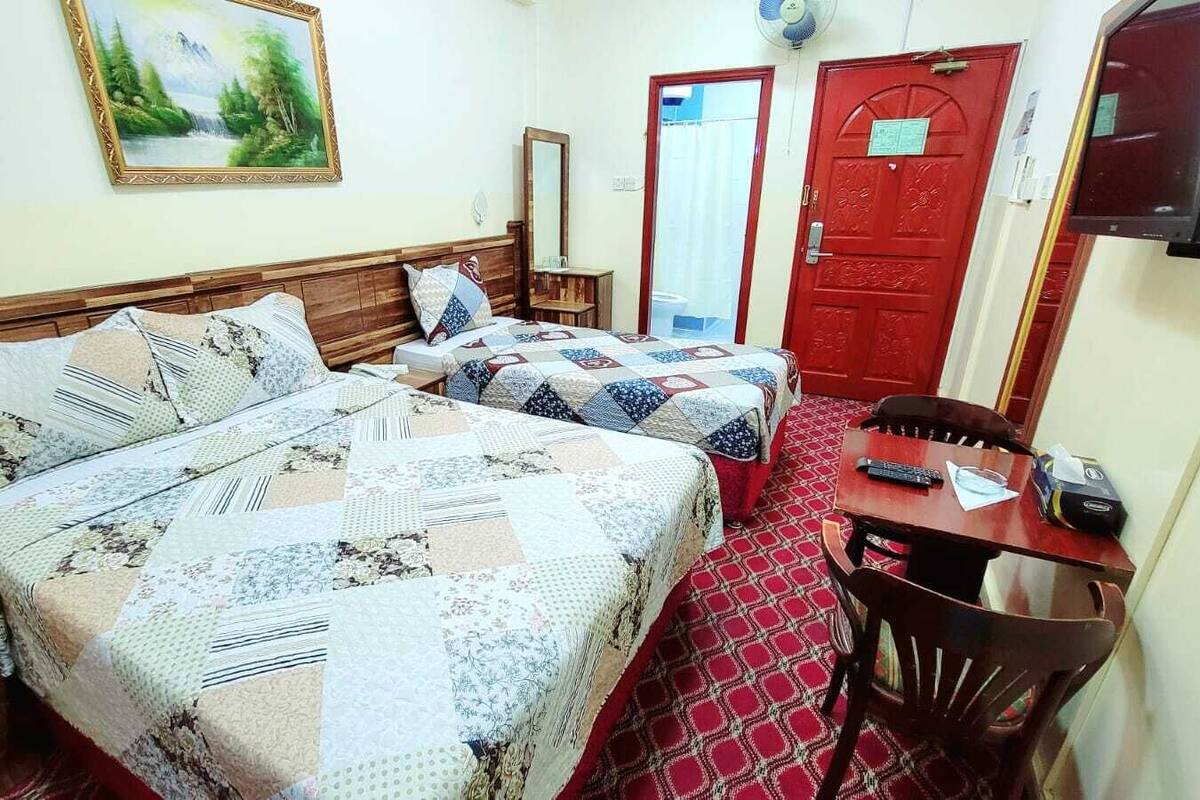 A hotel room with two beds, a wooden headboard, a red door, a small table with a chair, and a painting on the wall.