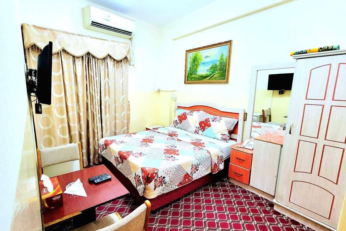 A hotel room with a bed, a desk with a chair, a wardrobe, and a painting on the wall.