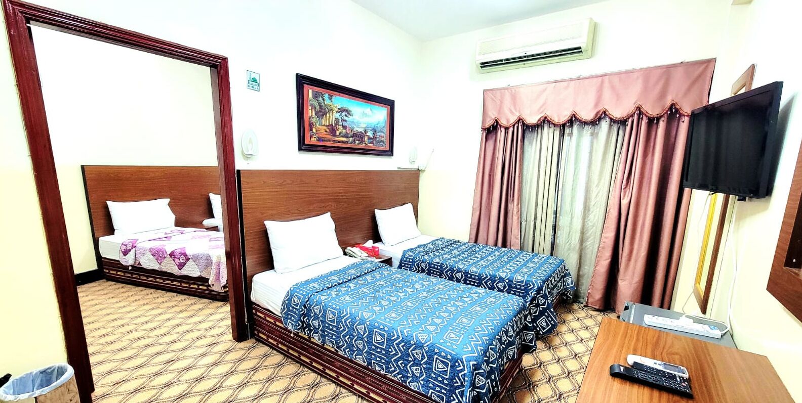 A hotel room with two beds, a wooden headboard, a painting on the wall, a TV, and a desk with a remote control.
