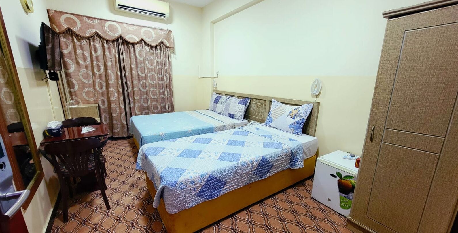 A hotel room with two beds, a desk, a chair, a wardrobe, and an air conditioning unit.