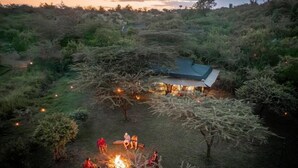 A rustic outdoor setting with a fire pit and a small building with a green roof surrounded by dense vegetation. - Basecamp Wilderness (Maasai Mara)