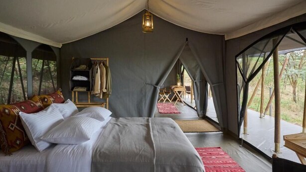 A tented accommodation room with a bed, a hanging light fixture, a small seating area, and a view of the outdoors. - Basecamp Wilderness (Maasai Mara)