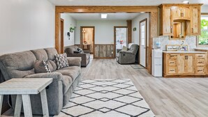 Ein modernes Wohnzimmer mit einer Couch, einem Sessel und einem Esstisch. Es gibt eine Küchenzeile mit Holzausbau und einem Kühlschrank.