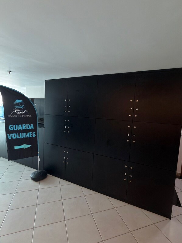 Un meuble de rangement noir doté de compartiments numérotés, une enseigne indiquant « GARDE-BAGAGES », et un logo pour « CASAS DE FERIAS ».