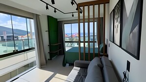 A modern living room with a sofa, a green accent wall, and a large window offering a view of buildings and a mountain. - LOFT BRISA 106 (Arraial do Cabo)