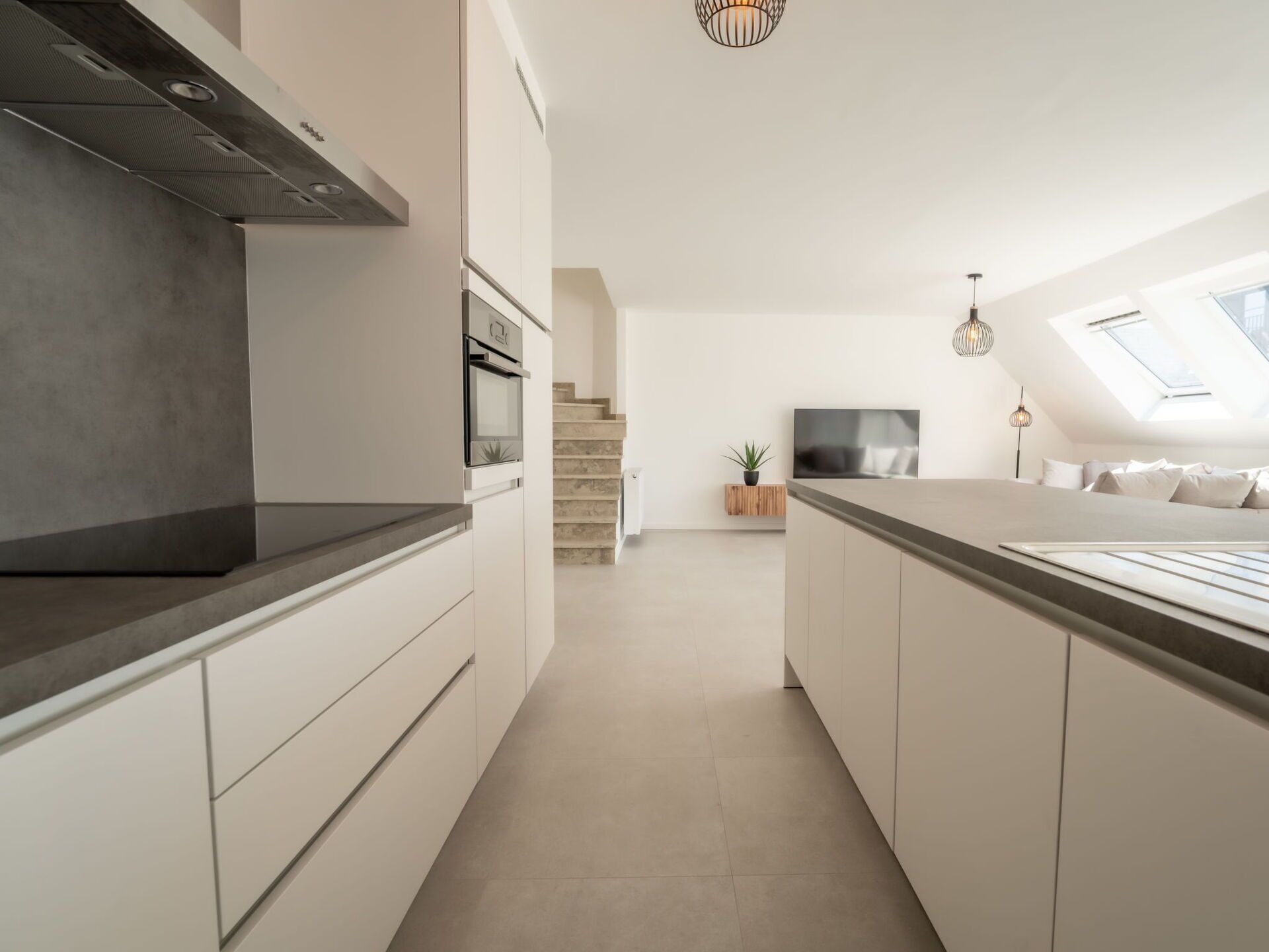 Une cuisine moderne dotée d’un îlot central, d’un four encastré et d’un escalier menant à une chambre à coucher aménagée en mezzanine, avec des fenêtres de toit.