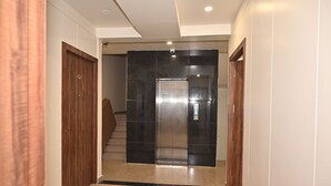 A hallway with a wooden door on the left, a black elevator in the center, and a wooden door on the right. - Atharv Kaashi - Behind Sigra Sports Stadium (Varanasi)