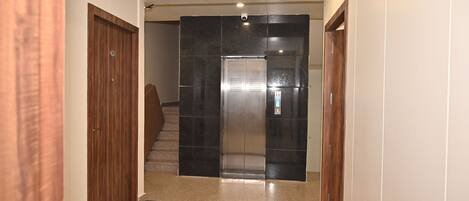 Un pasillo con una puerta de madera a la izquierda, un ascensor negro en el centro y otra puerta de madera a la derecha.