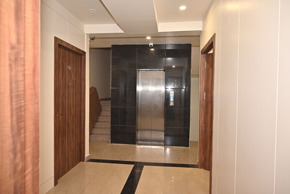 A hallway with a wooden door on the left, a black elevator in the center, and a wooden door on the right. - Atharv Kaashi - Behind Sigra Sports Stadium (Varanasi)