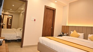 A hotel room with a large bed, a wooden door, a bedside table with a lamp, and a wall-mounted air conditioning unit. - Atharv Kaashi - Behind Sigra Sports Stadium (Varanasi)