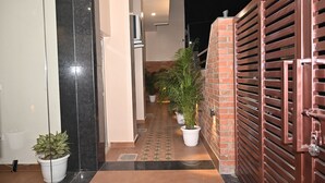 A corridor with tiled flooring, potted plants, and a brick wall with a metal gate. - Atharv Kaashi - Behind Sigra Sports Stadium (Varanasi)