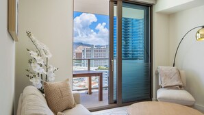 A modern living room with a sofa, a coffee table, and a view of a cityscape through a sliding glass door. - Modern 2BR at Sky Ala Moana – City & Mtn Views w/ AC/Pool/Gym/1 Parking (Honolulu)