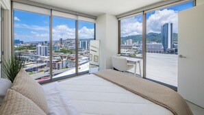 A modern bedroom with a large bed, a desk, and a view of a cityscape through floor-to-ceiling windows. - Modern 2BR at Sky Ala Moana – City & Mtn Views w/ AC/Pool/Gym/1 Parking (Honolulu)