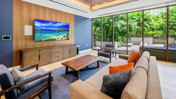 A modern living room with a large TV, comfortable seating, and a view of a lush garden through large windows. - Modern 2BR at Sky Ala Moana – City & Mtn Views w/ AC/Pool/Gym/1 Parking (Honolulu)