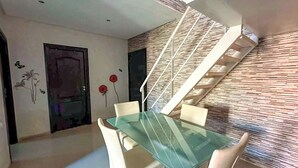 Een moderne eetruimte met een glazen tafel en witte stoelen, een trap met een gestructureerde wand, en een deur met bloemkunst.