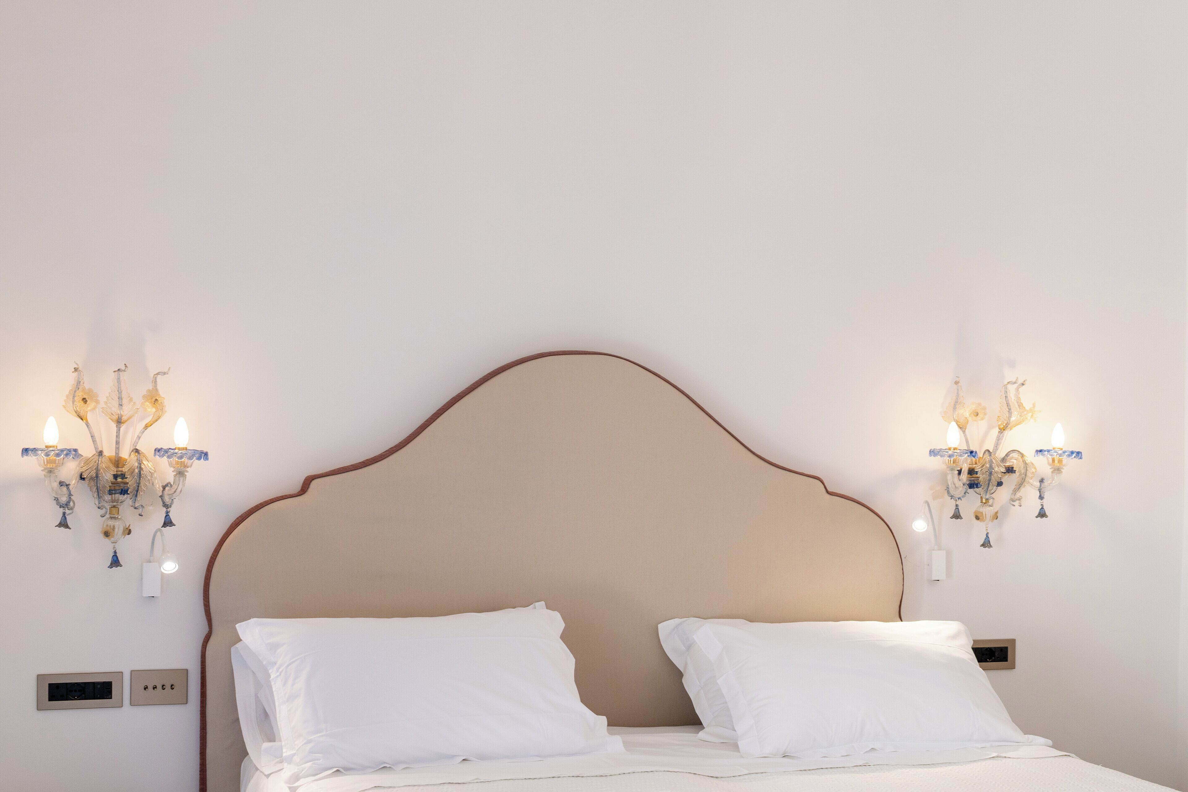 Camera da letto con una testiera beige, biancheria da letto bianca e due lampadari a parete.