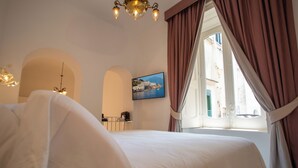A hotel room with a bed, a chandelier, a painting, and a view of buildings through the window.