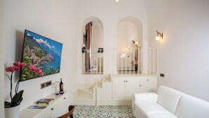 Una sala de estar moderna con un sofá blanco, un televisor montado en la pared, una escalera con barandilla y vista al mar a través de un arco.