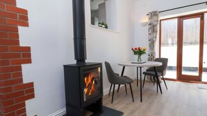 Un salon moderne avec un poêle à bois noir, une table de salle à manger ronde entourée de quatre chaises, un vase rempli de fleurs et une grande porte-fenêtre coulissante donnant sur un espace extérieur.