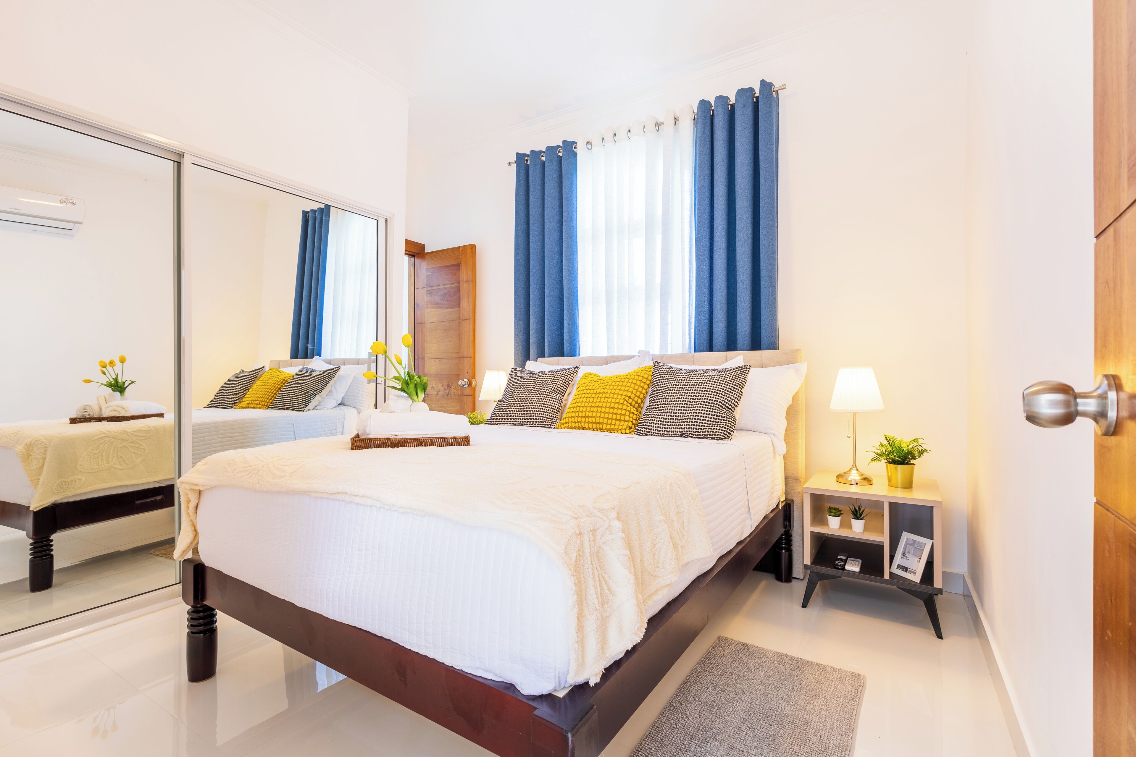 A modern bedroom with a large bed, a bedside table with a lamp, a small nightstand, and a mirror.