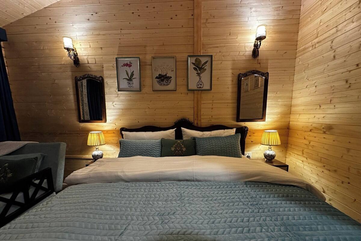 A wooden-paneled bedroom with a bed, two bedside lamps, and three framed botanical prints on the wall.