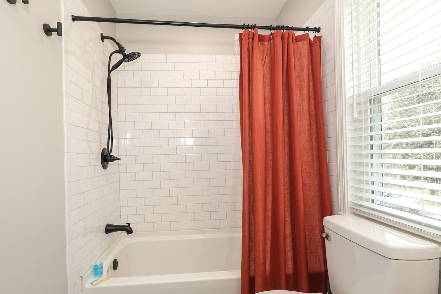 A bathroom with a white tiled shower, a black showerhead, a white bathtub, and a window with blinds.