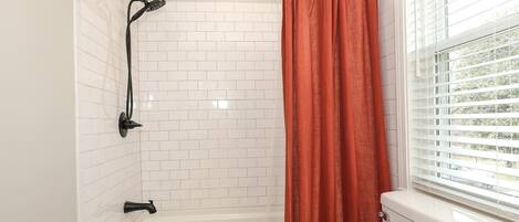 A bathroom with a white tiled shower, a black showerhead, a white bathtub, and a window with blinds.
