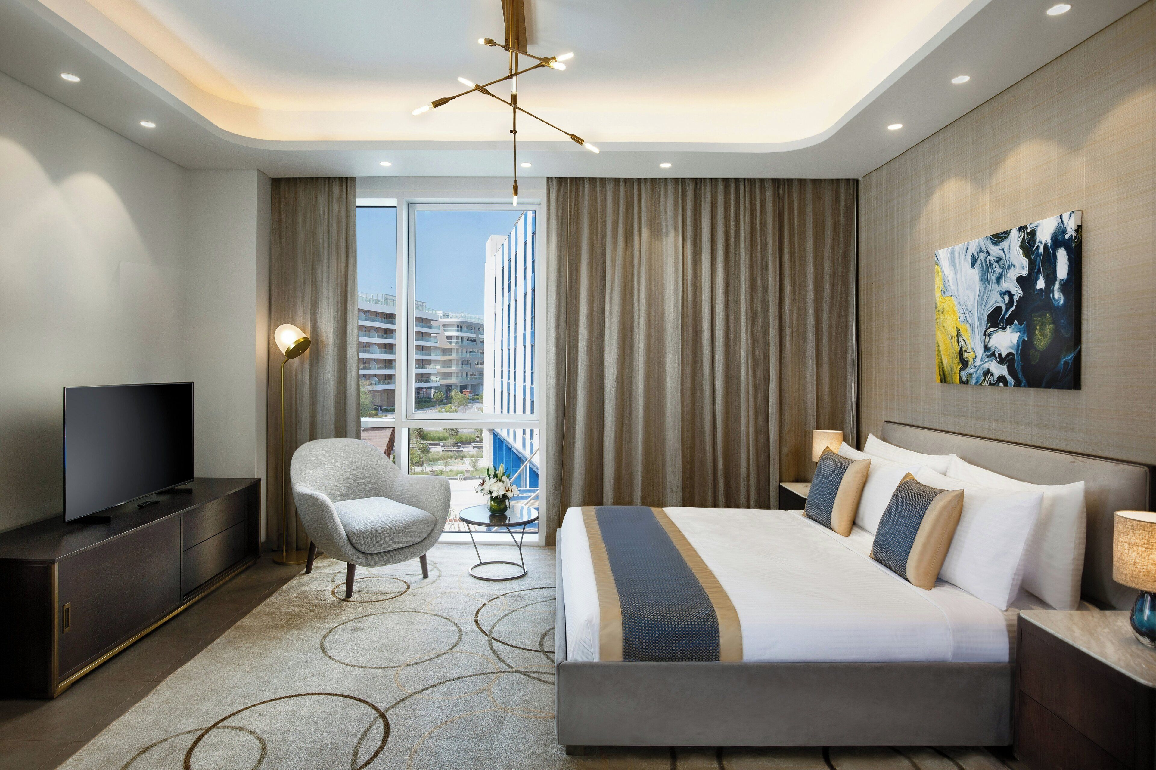 Quarto de hotel moderno com uma cama grande, televisão de tela plana, área de estar com uma poltrona, uma pequena mesa com um vaso de flores e vista da cidade por grandes janelas.
