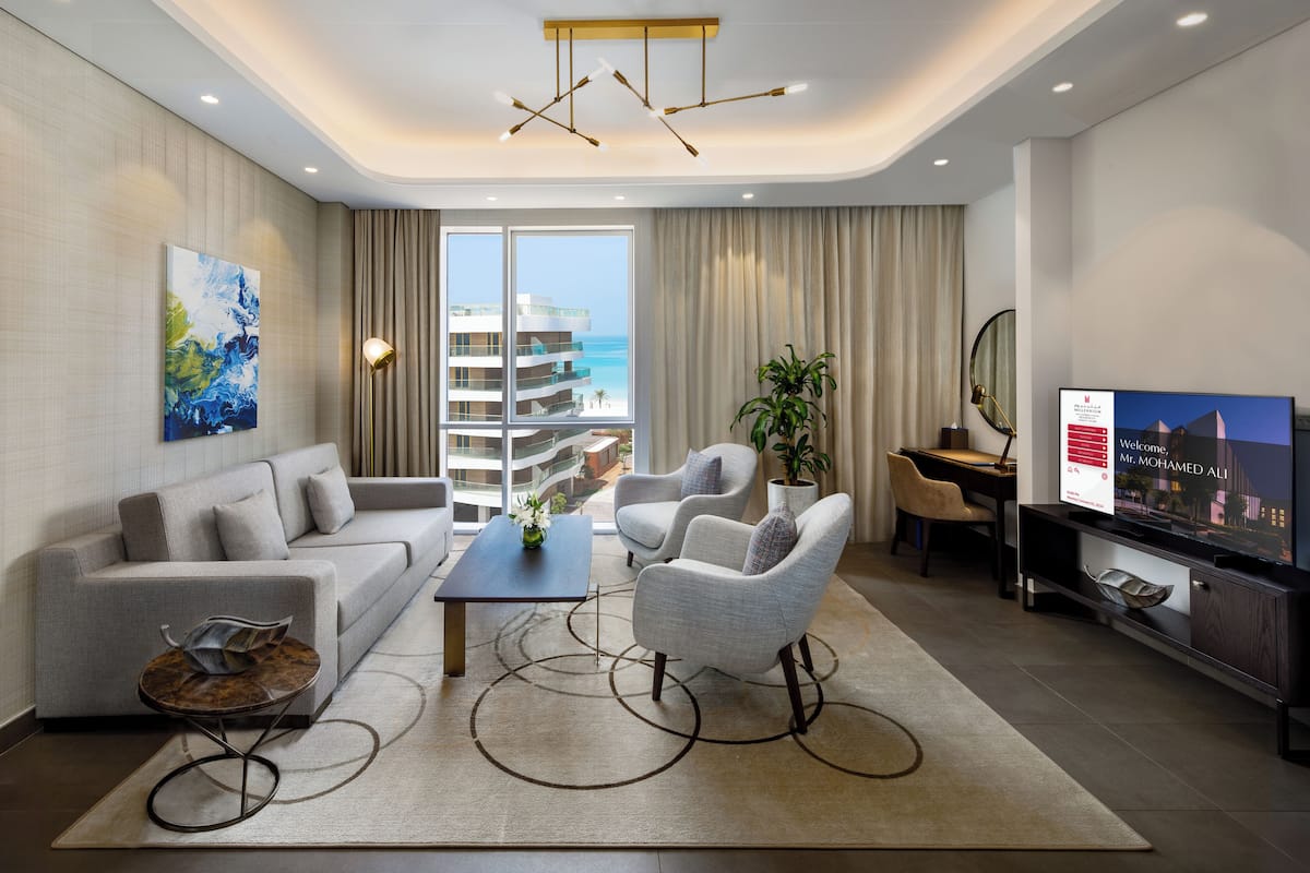 Una sala moderna con un sofá, sillones, una mesa de centro y un televisor. Hay un ventanal con vistas a un edificio y al mar.