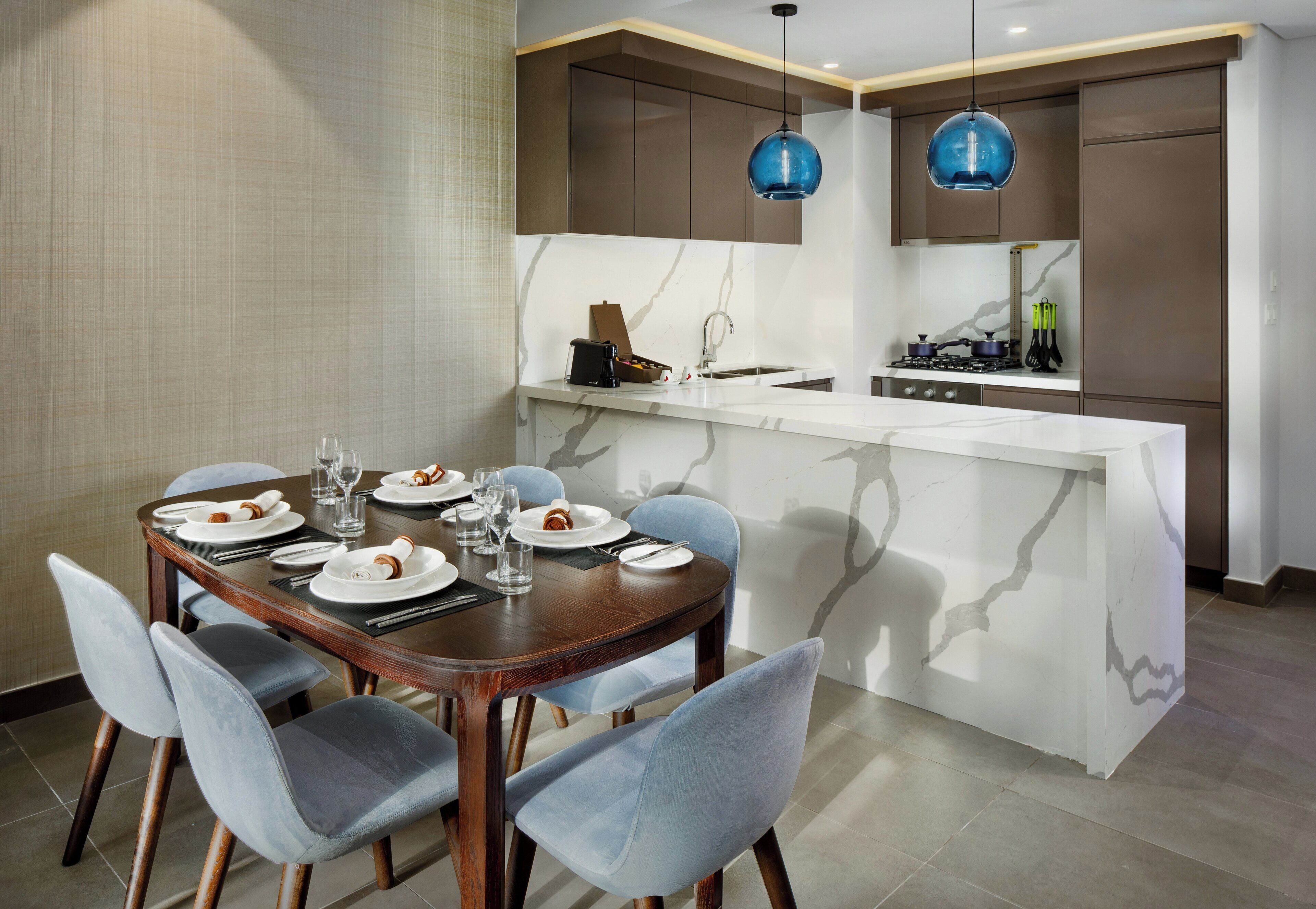 A modern kitchen with a marble island, dark wooden cabinets, and blue pendant lights.