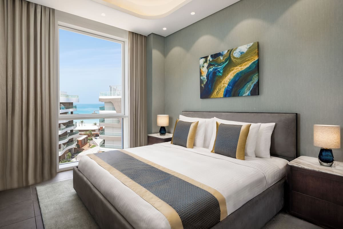 Una habitación de hotel con una cama grande, dos mesitas de noche, un buró, una ventana con cortinas y un cuadro en la pared.