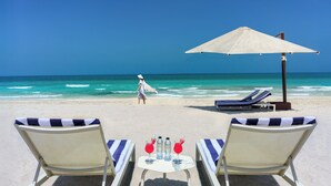 Blick auf einen Strand mit einem Tisch für zwei Personen, zwei Liegestühlen und einem Sonnenschirm.