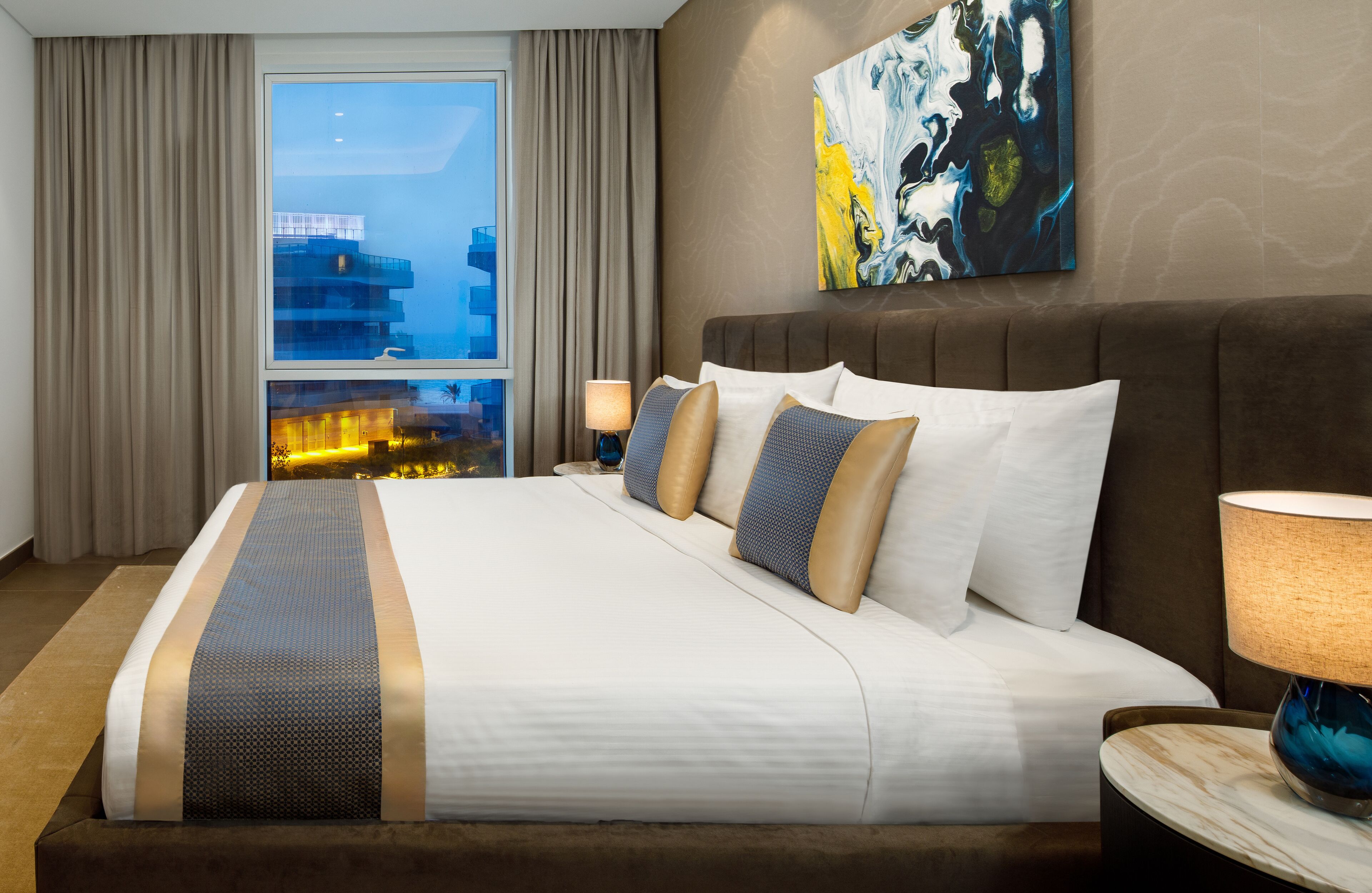 Habitación de hotel con una cama grande, vista panorámica de la ciudad a través de una ventana y un cuadro abstracto moderno en la pared.