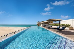 A swimming pool with a view of the beach, lounge chairs, and a covered seating area.