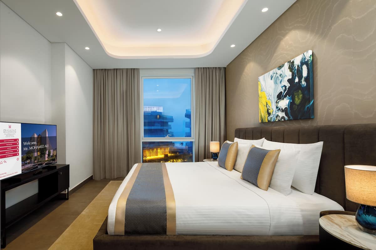 Una habitación de hotel moderna con una cama grande, un televisor de pantalla plana, una mesita de noche y vistas a la ciudad desde la ventana.