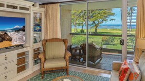 A living room with a television, a wicker chair, a sofa, and a view of a balcony with a lawn and palm trees.