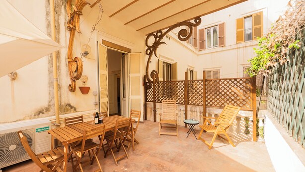 Une cour avec une table et des chaises en bois, une petite table ronde avec une bouteille, et une clôture décorative en métal.