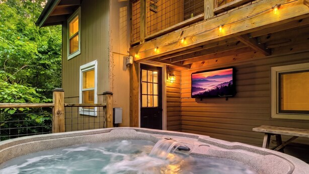 A hot tub with a wooden deck, a wooden house with a TV on the wall, and a door with a window.