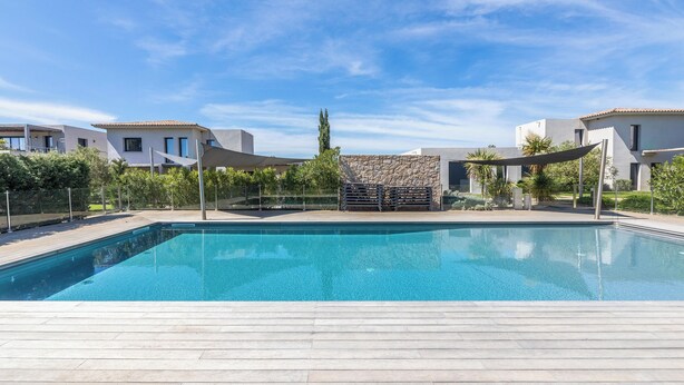 Una piscina rectangular con una terraza de madera en primer plano, rodeada de edificios modernos, un muro de piedra y follaje al fondo.