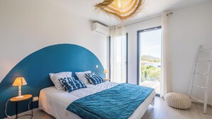 Un dormitorio con una pared azul curva, una cama con ropa de cama blanca y azul, una lámpara de noche, una ventana con vistas y una escalera apoyada en la pared.