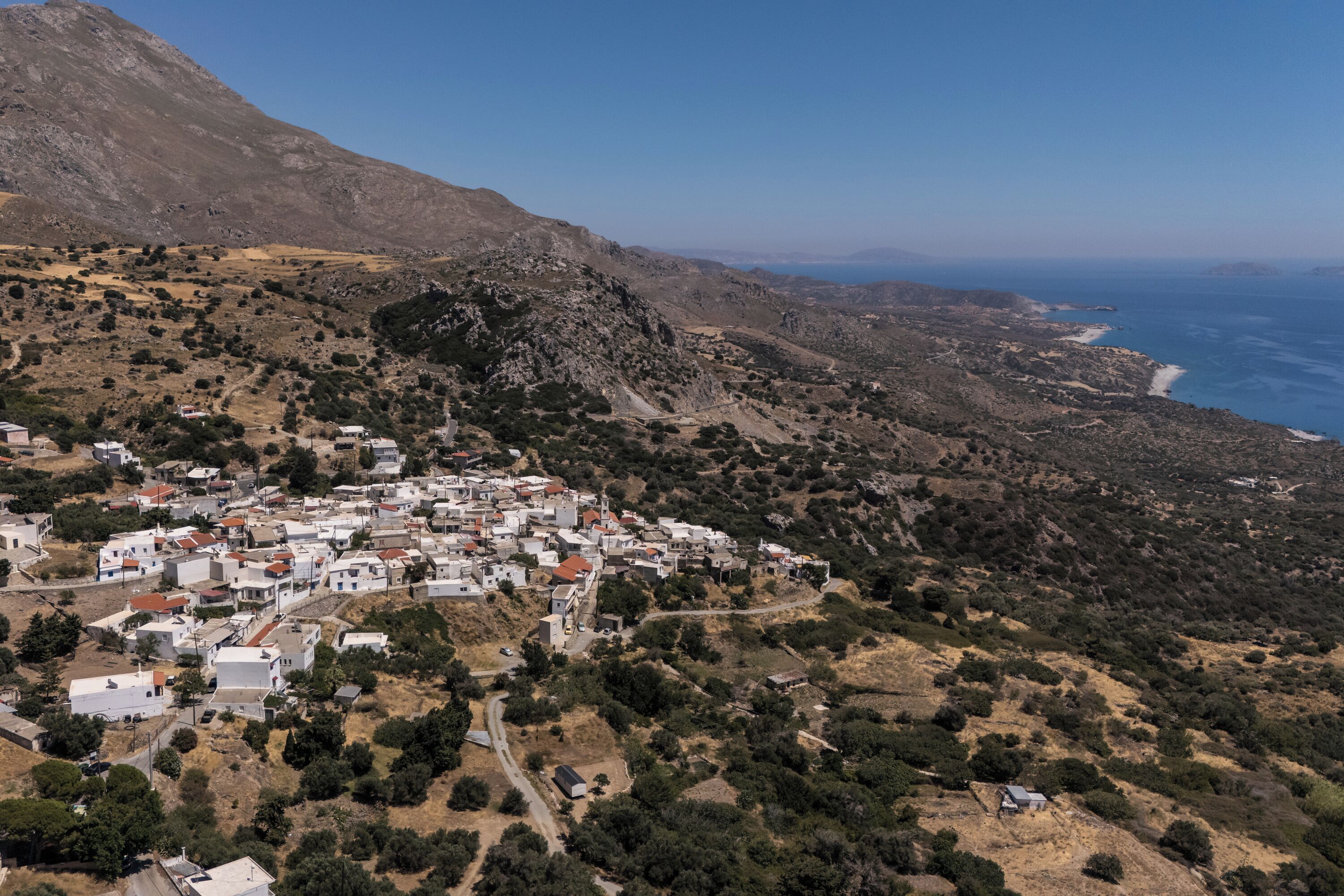 A coastal village with white buildings, a mountainous backdrop, and a view of the sea.