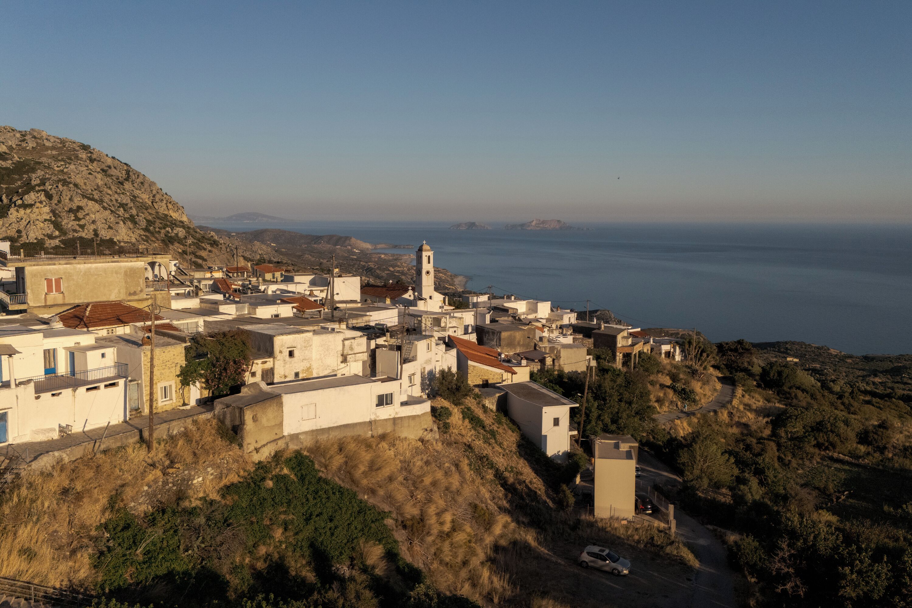 A coastal village with white buildings, a prominent church with a bell tower, and a road leading down a hillside.