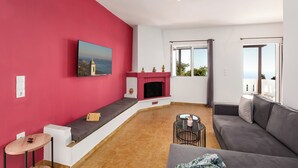 A living room with a red wall, a fireplace, a sofa, a table, and a balcony with a view. - Libyan Horizon Seaview (Rethymno)