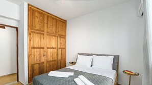 A neatly made bed with a quilted grey bedspread and two rolled white towels, a wooden wardrobe, and a small bedside table with a book and a lamp. - Libyan Horizon Seaview (Rethymno)